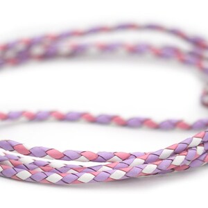 May include: A braided cord in shades of lavender, pink, and white. The cord is made of interwoven strands, creating a textured pattern. The cord is likely used for jewelry or crafting.