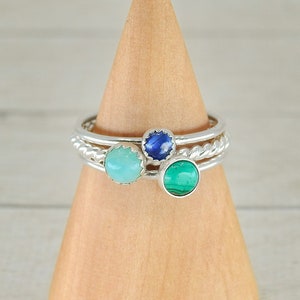 May include: Three silver rings with different colored gemstones. The rings are stacked on a wooden display stand. The top ring has a blue gemstone, the middle ring has a light blue gemstone, and the bottom ring has a green gemstone.