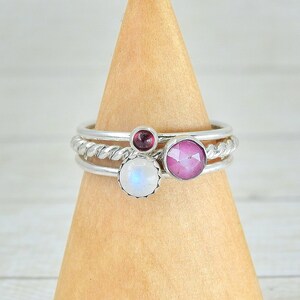 May include: Three silver rings with gemstones. The top ring has a pink gemstone, the middle ring has a white gemstone, and the bottom ring has a red gemstone. The rings are on a wooden display stand.