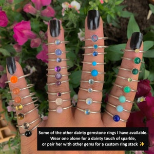 May include: A hand model wearing a variety of silver rings with different colored gemstones. The rings are stacked on each other and feature a variety of colors, including orange, red, purple, blue, green, and brown. The text "Some of the other dainty gemstone rings I have available. Wear one alone for a dainty touch of sparkle, or pair her with other gems for a custom ring stack ✨" is visible at the bottom of the image.