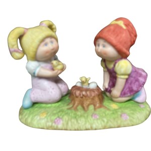 May include: Two porcelain dolls, one with blonde hair and a pink dress, the other with red hair and a purple dress, are kneeling beside a bird's nest with a small yellow chick inside.