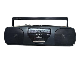 Op de afbeelding: Een zwarte draagbare Sony-radio met cassettespeler, voorzien van een handvat aan de bovenzijde, twee ronde luidsprekers aan de zijkanten, een centrale analoge radio-afstemming en een cassettecompartiment met het Sony-logo op de voorzijde.