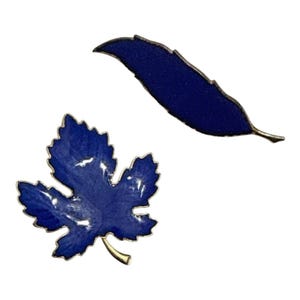May include: Two enamel pins shaped like leaves with a deep blue finish and metallic silver-toned edges. One pin is shaped like a broad, five-pointed maple leaf with a short stem, while the other is a slender, elongated leaf with a pointed tip.