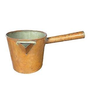 May include: A copper pot with a long, cylindrical handle and a triangular pouring spout. The exterior has a warm, metallic copper finish with visible signs of wear, while the interior features a light, silvery-grey metallic lining.