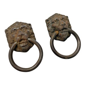 May include: Two antique bronze lion head door knockers. Each knocker features a detailed lion's head with a ring handle in its mouth. The bronze has a weathered, aged appearance, suggesting vintage craftsmanship. Ideal for adding a touch of classic elegance to any door.