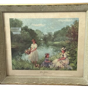 May include: Framed vintage print featuring a pastoral scene with three women near a body of water. The print is in a gold-colored frame with a textured border. The artwork depicts women in white dresses and hats, with one holding flowers.