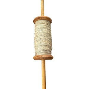 May include: A wooden kite spool with a light-colored string wound around a central wooden cylinder. The spool has a wooden handle extending from the top and bottom, creating a T-shape. The wood has a natural, light brown color.