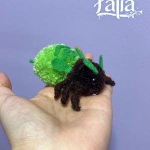 May include: A handmade plush insect toy with a green pom-pom body and brown legs and head. The toy has green felt accents and a black eye. The word "Palia" is written in white script in the upper right corner.