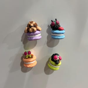 Könnte beinhalten: Vier farbenfrohe, skurrile Magnete in Form von Macarons. Jeder Magnet zeigt eine andere Kreatur: einen Hund, eine Fliege und zwei weitere insektenartige Figuren. Die Macarons sind in Lila, Blau, Orange und Gelb gehalten.