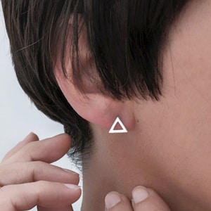 Puede incluir: Un pendiente de botón triangular plateado es visible en una oreja. El pendiente tiene una forma geométrica simple, un diseño minimalista. El cabello de la persona es oscuro y el tono de piel es claro.