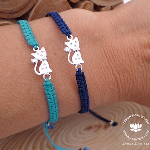 Może przedstawiać: Dwa regulowane bransoletki z srebrnymi zawieszkami w kształcie kota. Bransoletki są wykonane z turkusowego i granatowego sznurka. Zawieszki są ozdobione małymi kropkami. Bransoletki są zawiązane suwakowym węzłem.