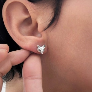 Puede incluir: Pendientes de plata con cabeza de zorro. Los pendientes son pequeños y delicados, y la cabeza de zorro es detallada y realista.