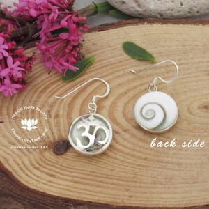 May include: Sterling silver earrings with a white shell on the back and a silver Om symbol on the front. The earrings are on a wooden surface with pink flowers.