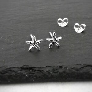 Puede incluir: Un par de pendientes de estrella de mar de plata. Los pendientes son pequeños y delicados, con un acabado texturizado.