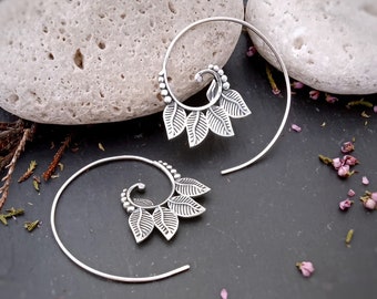 Op de afbeelding: Een paar zilveren spiraalvormige oorbellen met een bladontwerp. De oorbellen zijn gemaakt van een dunne draad die in een spiraalvorm is gedraaid. De bladeren zijn aan de spiraal bevestigd en zijn in een waaier vorm gerangschikt. De oorbellen zijn een delicaat en elegant sieraad.