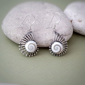 May include: A pair of silver-toned earrings featuring a spiral design with a white center, resembling a seashell. The earrings have a detailed, textured appearance with a hook closure. The earrings are displayed against a soft green background.