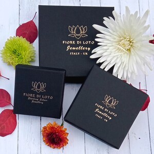 May include: A collection of black jewellery boxes, each with a gold lotus flower logo and text reading "FIORE DI LOTO Jewellery ITALY - UK". One box also states "Sterling Silver Jewellery". The boxes are surrounded by flowers.
