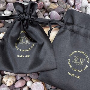 Puede incluir: Dos bolsas negras de sat&eacute;n con cord&oacute;n, texto dorado y logotipo de flor de loto. Las bolsas est&aacute;n etiquetadas como "DESIGN FIORE DI LOTO, ETHNIC, VINTAGE, CHIC, ITALY - UK". Las bolsas est&aacute;n sobre un lecho de piedras peque&ntilde;as, lisas y multicolores.