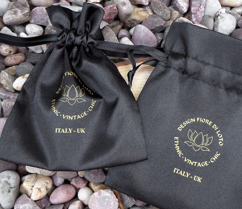 Puede incluir: Dos bolsas negras de sat&eacute;n con cord&oacute;n, texto dorado y logotipo de flor de loto. Las bolsas est&aacute;n etiquetadas como "DESIGN FIORE DI LOTO, ETHNIC, VINTAGE, CHIC, ITALY - UK". Las bolsas est&aacute;n sobre un lecho de piedras peque&ntilde;as, lisas y multicolores.