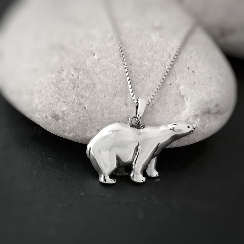 Puede incluir: Un colgante plateado de oso polar en una delicada cadena. El colgante es una escultura tridimensional detallada de un oso polar. El collar se muestra sobre un fondo borroso y neutro, destacando el dise&ntilde;o y la artesan&iacute;a de la joya.