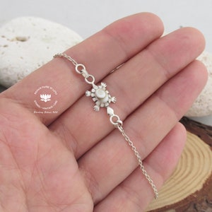 May include: A silver bracelet with a turtle pendant, featuring a white and beige centre. The chain is delicate, held in a hand. The text "DESIGN FIORE DI LOTO" and "Sterling Silver 925" are visible on the image.