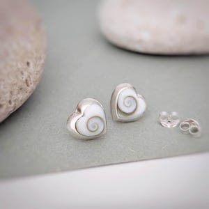 May include: Heart-shaped stud earrings with a silver frame and a white, spiral design. The earrings are presented with earring backs on a grey surface. The earrings are likely fashion jewellery.