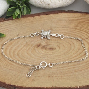 May include: A silver bracelet with a turtle charm. The charm has a white and beige shell. The bracelet has a delicate chain and an adjustable clasp. The turtle charm is the focal point of this jewellery piece.