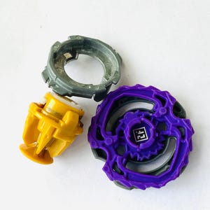 K&ouml;nnte beinhalten: Drei farbenfrohe Spielzeugkomponenten: ein grauer Ring, eine gelbe Basis und ein lila Oberteil mit einem QR-Code. Die Spielzeuge sind zum Drehen und K&auml;mpfen konzipiert. Die Komponenten bestehen aus Kunststoff.