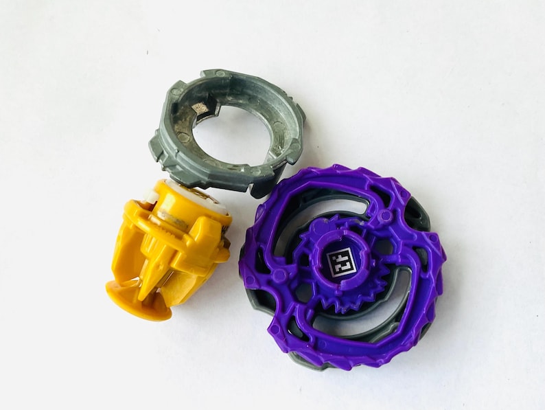 K&ouml;nnte beinhalten: Drei farbenfrohe Spielzeugkomponenten: ein grauer Ring, eine gelbe Basis und ein lila Oberteil mit einem QR-Code. Die Spielzeuge sind zum Drehen und K&auml;mpfen konzipiert. Die Komponenten bestehen aus Kunststoff.