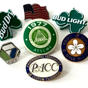 May include: A collection of seven vintage enamel pins. The pins feature various designs, including a Budweiser logo, an American flag, a Bud Light logo, a union logo, a flower, and a professional association logo.
