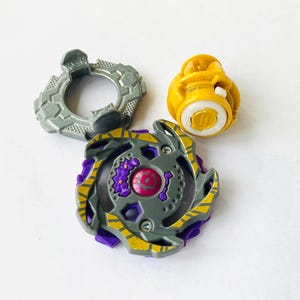 K&ouml;nnte beinhalten: Dreiteiliges Spielzeugset mit einem grauen, lila und gelben Kreisel. Der Kreisel hat ein kreisf&ouml;rmiges Design mit einem zentralen rosa Element. Ein grauer Ring und eine gelbe Komponente sind ebenfalls enthalten.