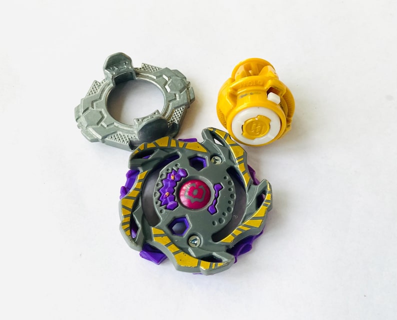 K&ouml;nnte beinhalten: Dreiteiliges Spielzeugset mit einem grauen, lila und gelben Kreisel. Der Kreisel hat ein kreisf&ouml;rmiges Design mit einem zentralen rosa Element. Ein grauer Ring und eine gelbe Komponente sind ebenfalls enthalten.