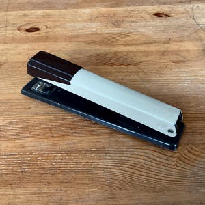 May include: A vintage stapler with a black base and a cream-colored body. The top portion is dark brown. The stapler is resting on a wooden surface with visible wood grain.