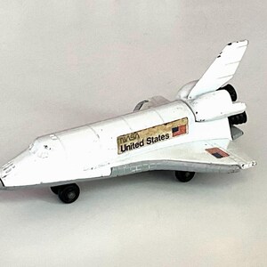 May include: A white toy space shuttle with black wheels and a grey undercarriage. The side features the NASA logo, the text United States, and two small American flag decals. The rear section includes black engine thrusters and a vertical stabilizer.