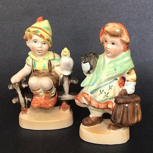 May include: Two porcelain figurines of children. The boy is wearing a green and yellow hat and holding a bird. The girl is wearing a green and white dress and holding a basket. Both figurines are standing on a base.