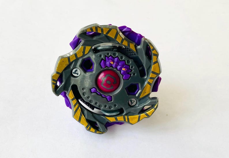 May include: A gray and purple toy with gold accents. The toy has a circular design with a central red emblem and a series of hexagonal and circular details. The edges are jagged and the overall appearance is mechanical.