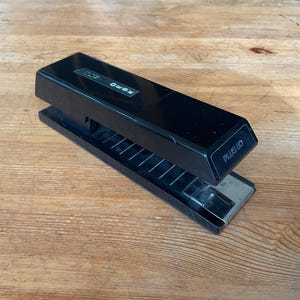 May include: Black stapler with the text "XEROX 300 PLUS GO" on a wooden surface. The stapler is open, revealing the staple compartment. The stapler is a rectangular shape.