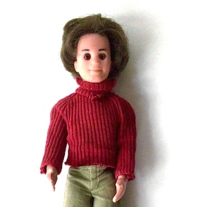 May include: A vintage doll with brown hair and a smiling face. The doll wears a red ribbed knit turtleneck sweater and olive green pants. The doll is standing against a white background.
