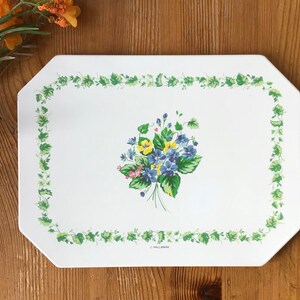 May include: White rectangular serving tray with a floral design. The tray features a border of green leaves and a central bouquet of blue, yellow, and pink flowers. The tray is on a wooden surface.