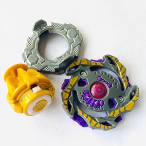 K&ouml;nnte beinhalten: Dreiteiliges Spielzeugset mit einem grau-gelben Oberteil, einer gelben Basis und einem grauen Ring. Das Oberteil hat lila und gelbe Akzente. Die Basis ist gelb mit wei&szlig;en und goldenen Details. Der Ring ist grau mit einem strukturierten Design.