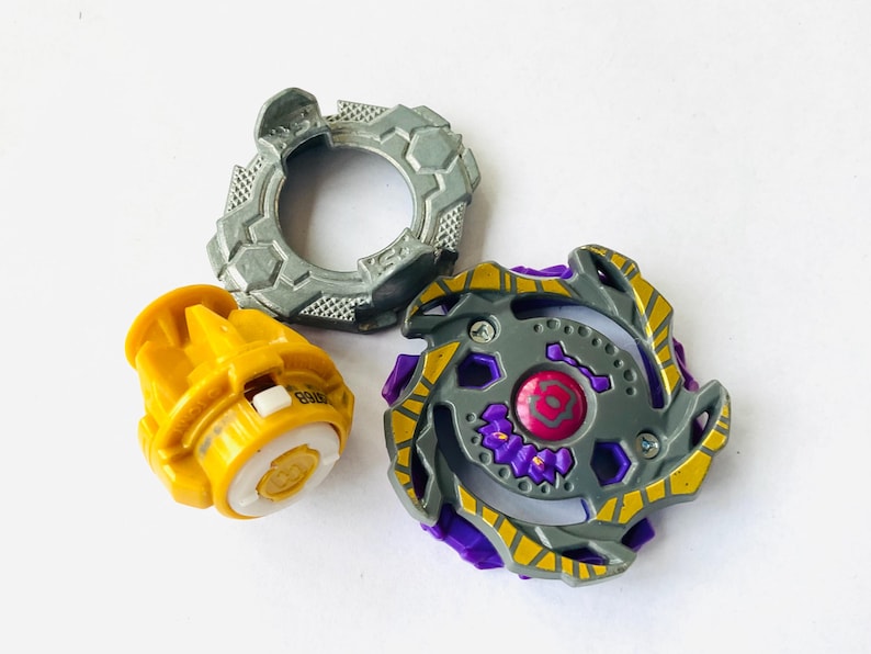 K&ouml;nnte beinhalten: Dreiteiliges Spielzeugset mit einem grau-gelben Oberteil, einer gelben Basis und einem grauen Ring. Das Oberteil hat lila und gelbe Akzente. Die Basis ist gelb mit wei&szlig;en und goldenen Details. Der Ring ist grau mit einem strukturierten Design.