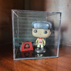 May include: A clear plastic display case with a red and white Christmas-themed box and a Funko Pop figure of a man wearing a brown jacket, a red shirt with the words "London Jets" on it, and a blue and white hat with a snowflake on it. The figure is standing on a black base. The text on the back of the case reads: "When you owe one thing, you've been to a party, you've seen time run, you've played pool, and you've given blood, but never thought life was to take an odd turn."