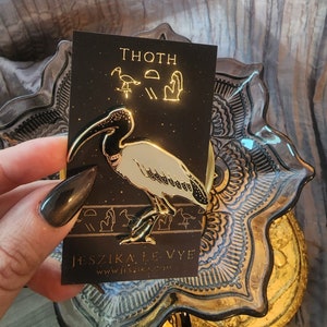 Puede incluir: Pin de esmalte dorado con un ibis blanco con marcas negras. El pin está en una tarjeta negra con el texto "THOTH" y "JESZIKA LE VYE" en letras doradas.