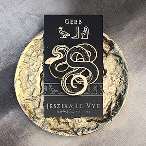 May include: A gold-coloured snake pin with a black background, featuring the name "GEBB" and hieroglyphic symbols. The pin is displayed on a textured, gold-coloured, round surface. The text "JESZIKA LE VYE" and website address are also visible.