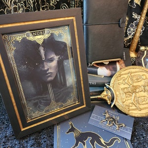 May include: A framed print of Anubis, the Egyptian god of the dead, with a black background and gold accents. The print is surrounded by a black frame with a gold trim. The image also includes a gold pin of Anubis, a black leather journal, and a gold-colored decorative object.