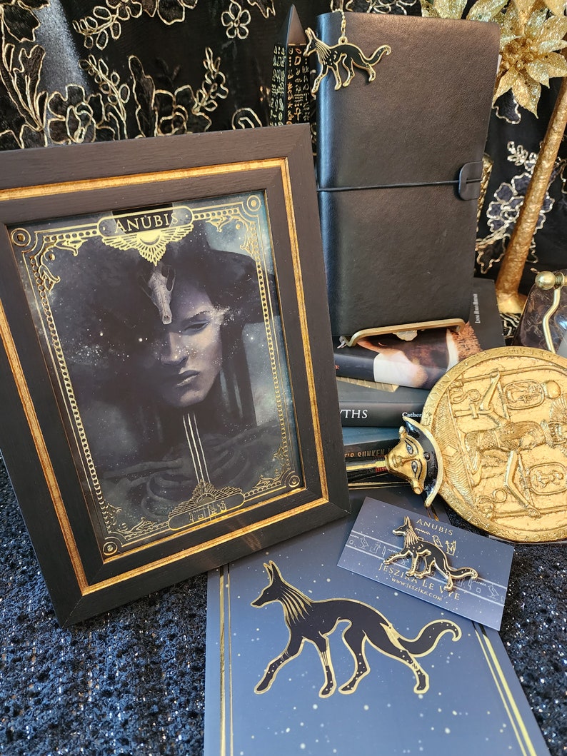 May include: A framed print of Anubis, the Egyptian god of the dead, with a black background and gold accents. The print is surrounded by a black frame with a gold trim. The image also includes a gold pin of Anubis, a black leather journal, and a gold-colored decorative object.