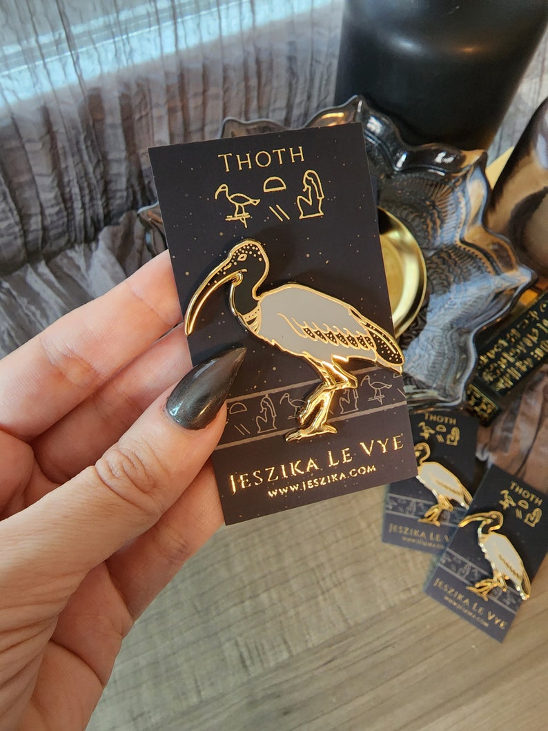 May include: Gold-colored enamel pin featuring a stylized ibis bird with black and white details. The pin is on a black card with the text "THOTH" and "JESZIKA LE VYE" in gold lettering. The card also features a small stylized ibis bird and hieroglyphs.