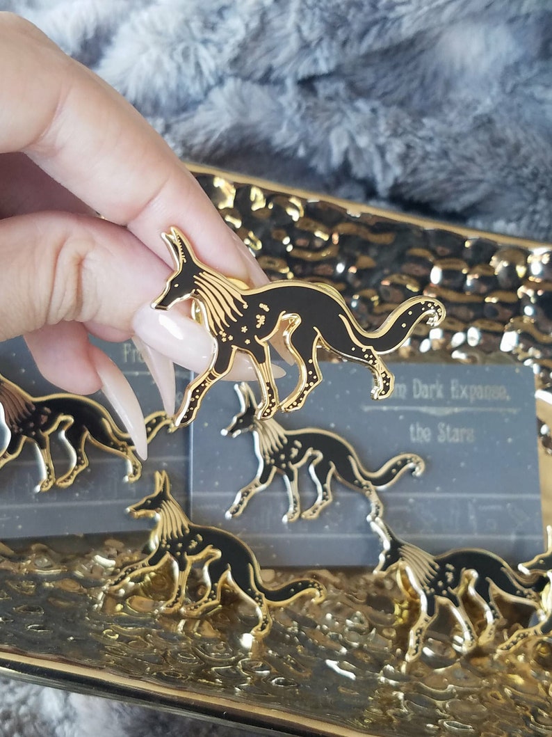 May include: A set of four gold-toned enamel pins featuring a stylized black wolf with a constellation design. The pins are arranged on a gold-colored surface. The text "In Dark Expanse, the Stars" is visible on a card behind the pins.