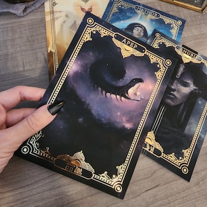May include: A set of tarot cards with gold accents and illustrations of Egyptian deities. The cards feature the names "Aton", "Hathor", "Apep", and "Anubis".