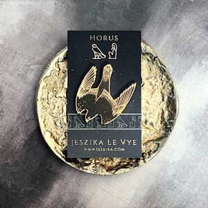 May include: A gold-colored enamel pin featuring a stylized depiction of the Egyptian god Horus, with the text "HORUS" and "JESZIKA LE VYE" and the website address "www.jeszika.com" on a black background.